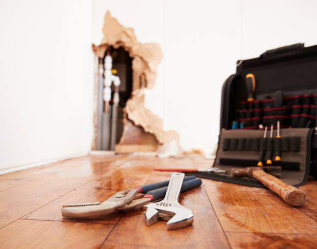 water damage prevention tips for flooring - repair tools in front on a wet wood floor with exposed plumbing in the background